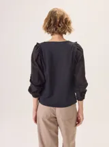 Camisa de color marrón con diseño combinado, presentando mangas abullonadas con volados en los hombros y cuerpo de algodón. Combina una textura de tela de camisa en mangas y delantero con una espalda de tejido elástico.