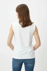 Remera de algodón blanca, de corte holgado y sin mangas, con cuello a la base y detalle de ojalillos metálicos sobre los hombros.