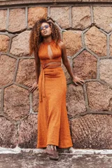 Pollera larga de tejido de punto color naranja con destellos brillantes, presenta un tajo frontal y una faja de ajuste en la cintura con lazo lateral.