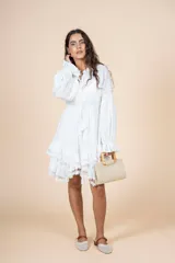 Vestido corto color blanco, de corte holgado con cintura elástica. Presenta escote en V con lazo de ajuste, mangas largas abullonadas con puños elásticos y falda con volados decorados con encaje en el ruedo.