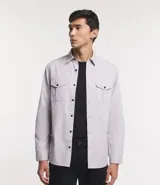 Camisa de manga larga para hombre, color gris claro, confeccionada en sarga de algodón. Presenta cuello clásico, cierre frontal de botones oscuros y dos bolsillos de parche con solapa y botón en el pecho.