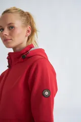 Campera de neopreno color rojo con capucha ajustable, cierre frontal completo y bolsillos laterales. Presenta puños ajustables con velcro y un parche circular decorativo en la manga izquierda.
