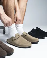 Zuecos cerrados al frente color negro, confeccionados en material símil gamuza. Presentan una tira ajustable con hebilla metálica en el empeine y una suela con detalle de corcho en el talón.