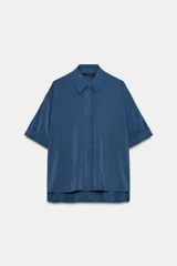 Camisa de corte fluido con cuello solapa y manga corta con dobladillo. Presenta un cierre frontal con botones ocultos bajo una tapeta y un diseño con bajo ligeramente más largo en la parte posterior.