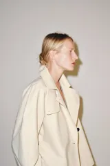 Trench corto color marfil de algodón, con cuello camisero, solapa cortaventos, mangas largas amplias con puños abotonados y cierre cruzado con un botón visible.