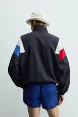 Chaqueta técnica de corte relajado con cuello subido y cierre frontal de cremallera bidireccional. Presenta un diseño color block en tonos azul marino, blanco, rojo y azul eléctrico. Incluye bolsillos frontales de vivo, puños y ruedo elásticos, y forro interior de red.