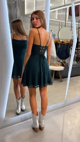 Vestido corto de fiesta color verde oscuro con acabado brillante y sutiles destellos. Presenta escote tipo cowl (caído), breteles finos y falda evasé con caída fluida.