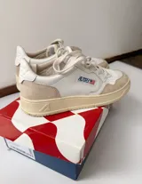 Championes Autry de diseño retro, color blanco con detalles en gamuza beige y suela de tono tostado, con cordones claros y logo lateral de la marca con la bandera de Estados Unidos.