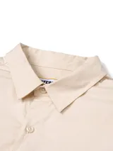 Camisa de manga larga color beige, estilo utilitario, con cuello clásico y cierre frontal de botones. Presenta tres bolsillos de carga en el pecho con solapas y cierre de velcro, además de un detalle de argolla metálica triangular y etiqueta con logo de la marca.