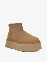 Bota corta tipo botineta de la marca UGG, color negro, con diseño de caña baja y plataforma elevada. Presenta una tira de agarre en color amarillo neón en la parte posterior y una suela de goma gruesa con textura acanalada.