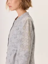 Camisa de algodón color gris claro, con cuello mao, cierre frontal de botones y mangas largas. Presenta un estampado sutil de hojas y flores, y detalles de bordado calado en los hombros.