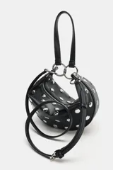 Bolso de mano tipo saca con diseño redondeado, confeccionado en material sintético color marrón oscuro. Presenta múltiples tachas metálicas doradas distribuidas por toda la superficie y un asa superior sujeta por anillas metálicas doradas. Cuenta con cierre superior mediante cremallera metálica.