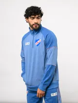 Buzo deportivo de medio cierre marca Umbro del Club Nacional de Football. Presenta un diseño en tonos azul y gris jaspeado, con cuello alto, mangas largas y detalles de costuras en contraste. Incluye el escudo del club y logo de la marca en el pecho.