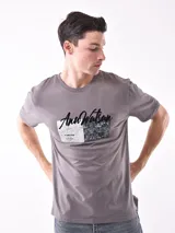 Remera de manga corta color gris topo, corte slim, con cuello redondo y un estampado gráfico en el pecho que combina texto manuscrito con un diseño abstracto en tonos oscuros.