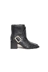 Bota corta de cuero color negro con textura efecto piel de serpiente, punta fina y tacón cuadrado medio. Presenta una tira cruzada sobre el empeine con una hebilla metálica dorada cuadrada en el lateral.