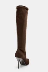 Bota alta de caña media en color marrón, confeccionada en material tipo gamuza. Presenta un diseño de punta fina, tacón alto y fino, y una tira decorativa con hebilla metálica en la parte superior de la caña. Incluye cierre lateral mediante cremallera.