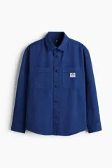 Sobrecamisa azul de lona gruesa de algodón, con cuello, tapeta con botones a presión y canesú en la espalda. Tiene mangas largas con botón a presión en los puños y dos bolsillos superiores de parche. El bajo es levemente redondeado y el corte es oversized.