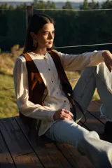 Camisa de color crudo con cuello redondo, volados sutiles en la pechera y botones frontales. Presenta mangas largas con puños sencillos y un diseño de corte recto.