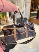 Bolso de viaje tipo duffel de cuero negro con detalles de refuerzo en gamuza y doble asa de mano.