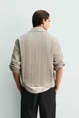 Chaqueta de punto con textura de trenzas verticales, color beige, cuello solapa y cierre frontal con cremallera.
