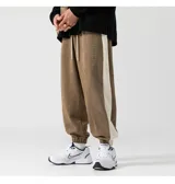 Pantalón tipo babucha de tela marrón tipo gamuza, con cintura elástica y cordón ajustable, puños ajustados en los tobillos y una franja lateral de color beige.