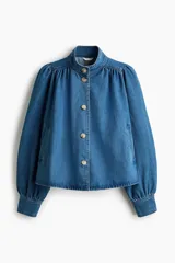 Campera de sarga de mezclilla azul, con cuello elevado, cierre frontal de botones y canesú fruncido. Tiene mangas largas abullonadas con puños ajustados y bolsillos laterales insertados.
