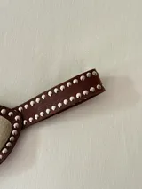 Cinto faja de cuero marrón con tachas metálicas plateadas en los bordes. Presenta una pieza central metálica con textura martillada y un dije en forma de gota con piedra natural.