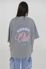 Remera color crema de corte holgado, con cuello redondo y estampado pequeño en el pecho con la frase 'Karma Club' en azul y rojo.