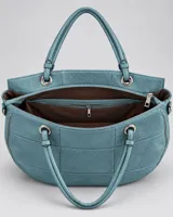 Cartera color celeste de diseño amplio y base redondeada, con textura granulada, costuras geométricas, doble asa de hombro y detalles metálicos con cierres.
