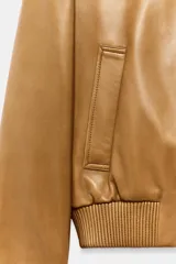 Campera bomber de cuero 100% color tostado de la ZARA WOMAN COLLECTION, con cuello redondo, manga larga, bolsillos delanteros ribeteados y cierre frontal con botones metálicos en relieve. Presenta elástico fruncido en el dobladillo inferior.