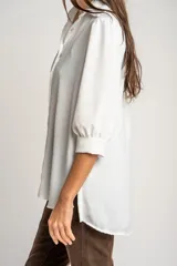 Blusa de corte oversize en color blanco, con cuello camisero, cierre frontal de botones y mangas tres cuartos con sutiles frunces en los hombros.