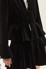Saco de terciopelo negro con cierre frontal mediante lazos, cuello mao, mangas largas con puños acampanados y detalle de volado en la cintura.