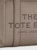 Cartera tipo tote de Marc Jacobs, confeccionada en cuero granulado color azul marino. Presenta el logo de la marca estampado en el frente, doble asa superior y correa ajustable y extraíble para llevar al hombro o cruzada.
