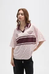 Remera tipo chomba de manga corta con cuello camisero, confeccionada en tejido con estampado de tablero ondulado en tonos rosado y blanco, con franja horizontal y detalles gráficos en color bordó.