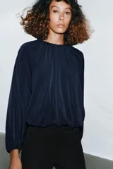 Top azul marino de tejido fluido con cuello redondo y manga larga abullonada con aberturas. Presenta una abertura en la espalda con tira del mismo tejido y cierre de ganchos metálicos.