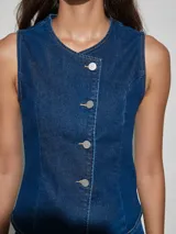 Chaleco de jean azul oscuro, sin mangas, con cuello redondo y cierre frontal de botones metálicos dispuestos en línea curva hacia el hombro izquierdo.
