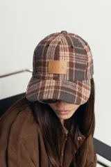 Gorra de estilo béisbol con estampado de cuadros tipo tartán en tonos marrones y grises. Presenta un parche frontal de cuero sintético con la marca grabada y cierre ajustable en la parte posterior.