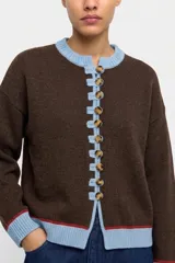 Saco tejido de punto en color marrón, con detalles en contraste color celeste en el cuello, puños y a lo largo de la botonera frontal. Presenta cierre con botones y una línea decorativa en color rojo en los bordes inferiores.