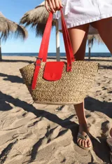 Bolso tipo canasto de paja con detalles en cuero rojo, incluyendo un bolsillo externo con solapa y cierre con botón a presión, y correa regulable.