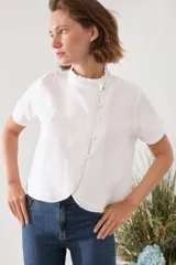 Blusa de lino blanca de manga corta y cuello alto con volado, con cierre asimétrico de botones dorados y costuras pespunteadas a contratono.