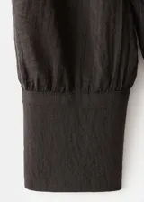 Blusa de color marrón oscuro con textura, cuello alto envolvente con detalle de fular integrado, manga larga con puños abotonados y diseño recto.