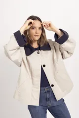 Chaqueta corta de color beige con cuello y puños vueltos en contraste de color azul marino, cierre frontal de botones oscuros y bolsillos ribeteados.