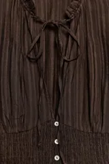Top de color marrón con textura acanalada, cuello redondo con lazada y escote en pico. Presenta mangas cortas abullonadas con terminación elástica, cuerpo con tejido tipo nido de abeja y cierre frontal con botones.