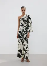 Mono largo de viscosa con diseño bicolor en negro y beige con estampado de hojas tropicales. Presenta un corte asimétrico de un solo hombro con manga larga holgada, pantalón de silueta recta y lazo ajustable en la cintura.