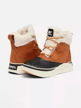 Bota de estilo pato Sorel, confeccionada en cuero impermeable color tostado con forro interior de borrego sintético. Presenta una carcasa de goma vulcanizada negra en la base, entresuela de plataforma en color crema y cierre con cordones.