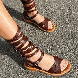 Sandalias planas de cuero color marrón, con tiras cruzadas en el empeine y múltiples tiras que se enrollan en la pierna, ajustables con hebillas metálicas.