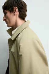 Trench de corte relajado confeccionado en tejido técnico italiano, color beige. Presenta cuello camisero, manga larga, bolsillos de vivo en la cadera y cierre frontal de botonadura simple con botones oscuros.