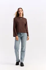 Remera de manga larga color beige con silueta relajada y mangas amplias. Presenta una estampa en la espalda con la frase 'Be kind to your mind' acompañada de ilustraciones de un cupido, flores y lazos en tono bordó.