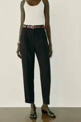 Pantalón tobillero de tiro medio, color beige, con pinzas delanteras y bolsillos frontales. Incluye un cinturón fino de cuero marrón con hebilla metálica dorada y cierre frontal con cremallera, botón interior y gancho.