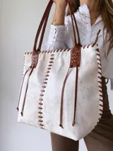 Cartera tipo tote de cuero vacuno con acabado texturizado en tono blanco hueso. Presenta detalles de costuras artesanales con tientos de cuero marrón en los bordes y correas de hombro a juego.